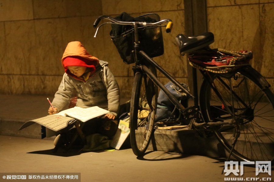 鄭州中原路京廣鐵路大橋下涵洞的馬路牙子邊，一位戴著紅色辣椒帽的小男孩趴在一個(gè)小馬扎上，上面鋪著一張車墊，在微弱的燈光下寫作業(yè)